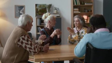 Huzurevinin rahat oturma odasında kağıt oynarken sohbet eden ve gülen bir grup çok ırklı yaşlı arkadaş.