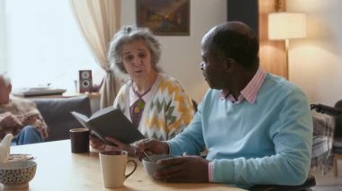 Kıdemli Afro-Amerikalı adam masada yemek yiyor ve yanında oturan beyaz kadınla konuşuyor ve bütün gün huzurevinde kitap okuyor.