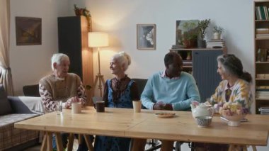 Bir grup çok ırklı yaşlı arkadaş, rahat oturma odasındaki ahşap masanın etrafında oturmuş, huzur evinde çay içerken sohbet ediyorlar.