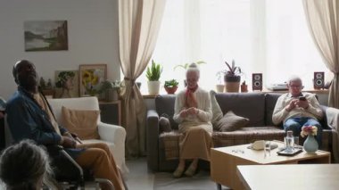 Beyaz kadın bakıcı mavi önlüklü siyah yaşlı adama tekerlekli sandalyede yardım ederken diğer yaşlılar da huzur evinde dinleniyor.