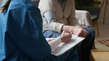 Huzurevinin oturma odasındaki kanepede oturan yaşlı gri saçlı adamı, kan basıncı testi sırasında not alan kadın doktorla konuşurken çek.