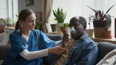 Kafkasyalı hemşire yaşlı siyahi bir adamın huzur evinde kanepede otururken nebulizörle ilaçlarını solumasına yardım ediyor.