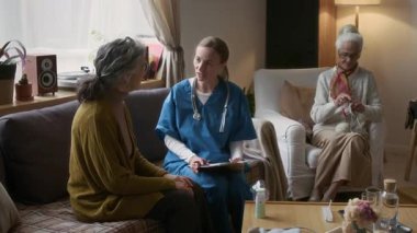 Huzur veren önlüklü hemşire, huzurevinin oturma odasında tıbbi konsültasyon sırasında kanepede otururken sağlık sorunlarından yakınıyor.