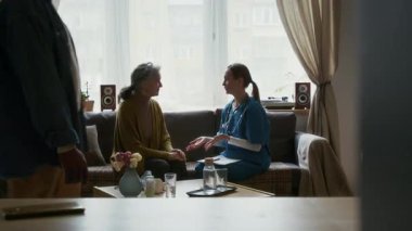 Kadın tıp asistanı, yaşlı bir kadınla kanepede oturuyor, gün boyunca konuşuyor ve ellerini tutuyor.