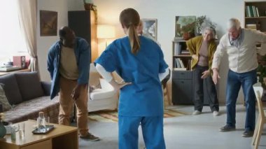 Mavi önlüklü bakıcı çeşitli yaşlılardan oluşan bir grup huzurevinin rahat oturma odasında esneme egzersizleri yapıyor.