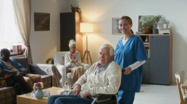 Mavi önlüklü, gülümseyen kadın bakıcı portresi huzurevinin rahat oturma odasında tekerlekli sandalyedeki yaşlı adamın arkasında duruyor, diğer yaşlılar arka planda oturuyor.