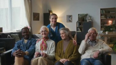 Çok ırklı mutlu yaşlıların toplu portresi ve önlüklü kadın bakıcı kamera önünde birlikte poz veriyor ve huzurevinin rahat oturma odasında gülümsüyor.