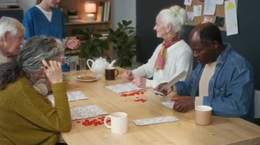 Bir grup çok ırklı kıdemli arkadaş ahşap masanın etrafında oturmuş tombala oynarken, mavi önlüklü kadın bakıcı yaşlı bir kadına gün boyunca bakım evinde su servisi yapıyor.