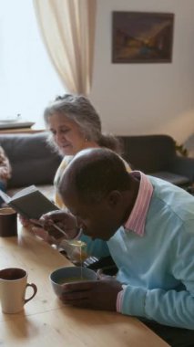Kafkasyalı yaşlı bir kadının kitap okuduğu ve Afrikalı Amerikalı bir arkadaşla emekliler evinde yemek yerken çekilmiş dikey bir fotoğrafı.
