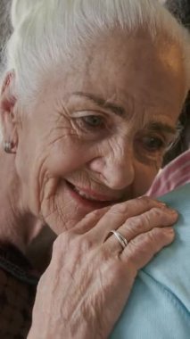 Beyaz saçlı, gülümseyen, başını partnerinin omzuna yaslamış ve huzur evindeki partide dans ederken kameraya bakan yaşlı bir kadının dikey portresi.