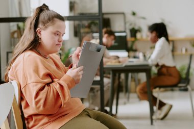 İş arkadaşları iş planlarını tartışırken elinde pano olan ve evrakları dolduran Down sendromlu kadın ofis çalışanının orta boy fotoğrafı.