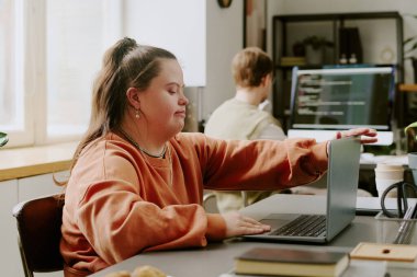 Down sendromlu bir programcının ofiste güneş ışınlarıyla aydınlatılan dizüstü bilgisayarını açarken orta boy görüntüsü.