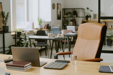 Üzerinde laptop ve ofis malzemeleri olan ahşap masa ve modern ofiste bej deri bir bilgisayar koltuğu.