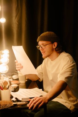 Genç adam soyunma odasında oturmuş parlak makyaj ışıkları altında belgeler okuyor, detaylara odaklanıyor, kozmetik ürünler ve fırçalarla çevrili, günlük giysiler giyiyor.