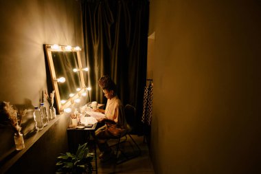 Soyunma masasında oturan, büyük ayna ve gösterişli ışıklarla hazırlanan, rahat soyunma odasında rahatlatıcı atmosfer yakalayan kişi.