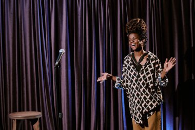 Afrika kökenli Amerikalı bir kadın sahnede stand-up komedisi yapmaktan zevk alıyor. Mikrofon ve tabure yanında hareketli bir ifadeyle.