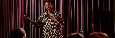 Afro-Amerikalı komedyen, elinde mikrofon, elinde stand-up gösterisi yaparken gülümseyen desenli tişört giyiyor.