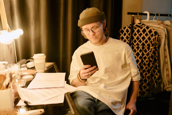 Man wearing green hat and glasses sitting near vanity table filled with papers and coffee cup while reading phone attentively