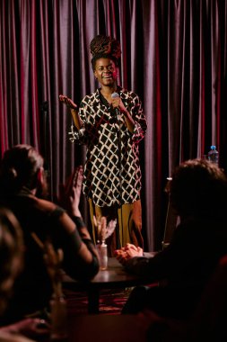 Sahnede perde ile süslenmiş, canlı bir atmosferde masalarda oturan seyircilerle etkileşim halinde olan Afro-Amerikalı bir kadın stand-up komedisi sergiliyor.