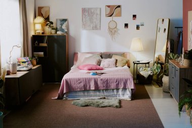 Ahşap mobilyalar, rahat halılar, duvarlarda soyut resimler ve saksılarda ev bitkisi ile donatılmış minimalist kız odasının geniş bir görüntüsü.