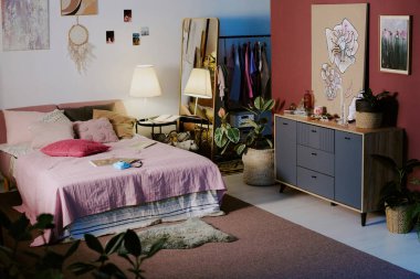 Minimalist yatak odası ahşap mobilyalar ve elbise askıları ve soyut resimler, posterler ve yeşil bitkilerle süslenmiş kadın giysileriyle dolu.