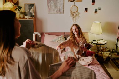 Arkadaşı yatakta mükemmel bir gece elbisesi seçerken elinde pijamaları olan iki askıyla tanınmaz bir esmerin yüksek açılı çekimi.