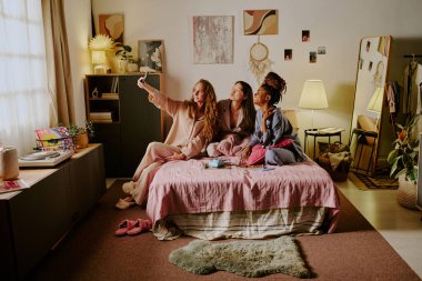 İpek pijamalı üç melez kadının yatıya kaldıkları sırada yatakta selfie çektikleri geniş açı.