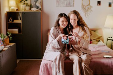 Pembe gül pijamalı iki arkadaş yatağın kenarına oturmuş ellerinde fotoğraf makinesiyle yeni fotoğraflara bakıyorlardı.