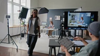 Beyaz gömlekli ve gri yelekli bir kadın el çırpma tahtası kullanırken, yönetmen de kameradan çiçeklerin, kitapların ve aydınlatma ekipmanlarının arasında rol alan genç bir kadın olarak parlak film setine bakıyordu.