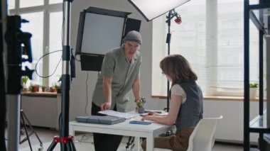 Erkek film yönetmeni beyaz masaya yaslanıyor, aktrisle detayları tartışıyor, büyük pencereli, parlak ışıklı stüdyoda profesyonel ışıklandırma ekipmanlarıyla çevrili.