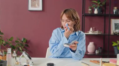 Açık mavi tişörtlü bir kadın, makyaj yaparken kameraya konuşuyor. Güzellik ürünleriyle dolu.