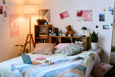 Yatak odasında, yastıkları ve kitap rafları ile birlikte, lamba ışığının altında güzelce yerleştirilmiş bir yatak. Çeşitli posterler ve renkli elementlerin yer aldığı dekore edilmiş duvarlar davetkar bir atmosfer yaratıyor.