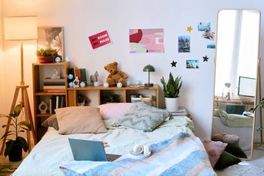 Sıcak ve davetkar bir atmosfer yaratan fotoğraflar, posterler ve bitkiler gibi kişisel dokunuşlarla dolu samimi bir yatak odası. Yataktaki dizüstü bilgisayar ve kulaklıklar rahatlama ve verimliliği öneriyor.