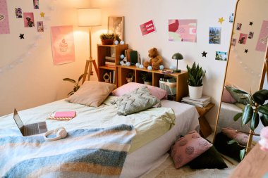 Parlak dekore edilmiş yatak odasında duvar sanatı renkli yastıklı rahat bir yatak ve kitaplarla dolu ahşap bir raf ve oyuncak ayı bulunuyor. Bilgisayarı ve kulaklığı olan bir çalışma alanı.