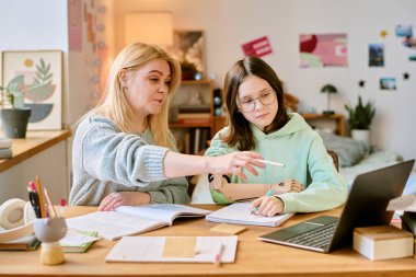 Konforlu ev ortamında protez eli olan genç bir öğrenciye rehberlik eden genç bir öğretmen. Masanın üzerinde kitaplar, duvarlarda posterler ve çalışma alanı sağlamanın yanında bitkiler.