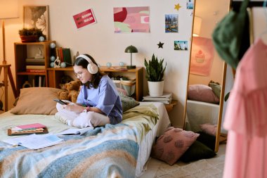 Protez eli olan, kulaklıkla çalışan, kitap ve dekorasyonla çevrili yatağında oturan, işine çok odaklanmış, bitki ve posterlerle süslenmiş bir çocuk.