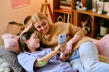 Sarışın anne ve protez elli esmer kızı kanepede oturmuş, tek bir akıllı telefonla gülümsüyorlar.
