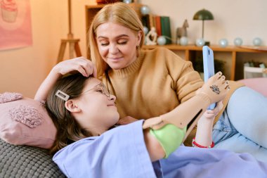 Annenin portresi evde kanepede otururken protez koluyla tablet kullanan çocuğa nazikçe bakıyor.