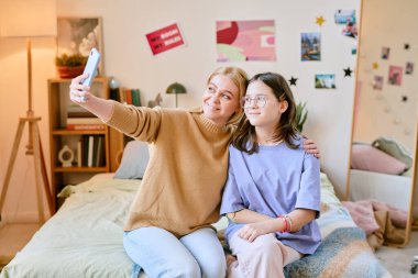 Sıcak yatak odasında selfie çekerken protez elli anne ve kızı gülümsüyor, posterler ve kitaplıklarla süslenmiş kişisel atmosferi yansıtıyor.