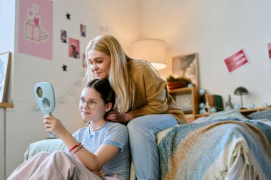 Protez kollu bir genç, omzuna yaslanmış bir ayna tutuyor. Hem gülümsüyor, hem de hoş bir dekorla rahat bir yatak odasında oturuyor. İç açıcı bir an yaşatıyor.