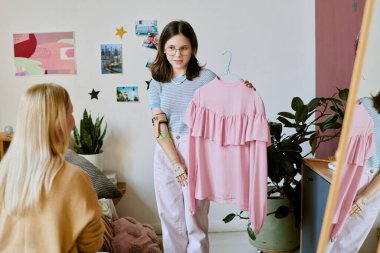 Protez kollu gülümseyen genç bir kız başka birine pembe elbisesini gösteriyor. Hoş, dekore edilmiş bir odada duvarlarda bitkiler ve sanat eserleri asılı.