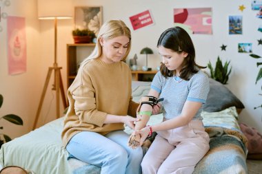 Beyaz bir anne, güzel kızına protez koluyla yardım ederken rahat bir yatak odasında, posterler, bitkiler ve yumuşak ışıklandırmayla süslenmiş bir odada sıcak bir atmosfer yaratıyor.