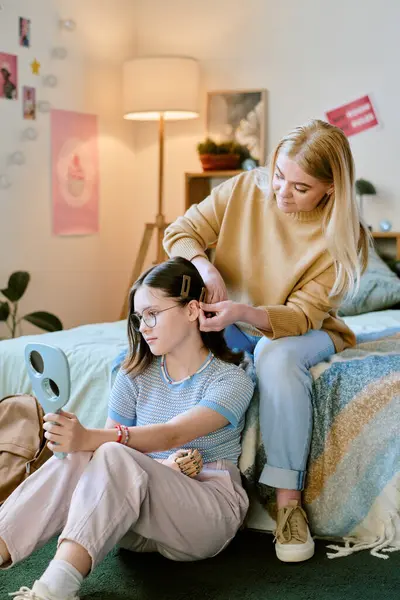 Yatakta oturan bir kadın, el aynası kullanırken genç kızların saçını düzeltiyor. Yumuşak ışıklandırma ve duvar fotoğraflarıyla dekore edilmiş bir oda, sade bir ortam yaratıyor.