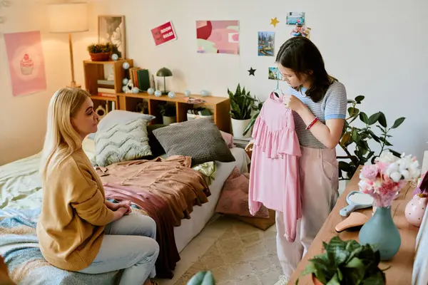 Anne ve kızı rahat bir yatak odasında pembe kıyafete hayran kalıyorlar. Arka planda çeşitli dekorasyonlar var. Sıcak bir atmosfer yaratıyorlar.