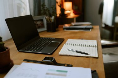 Sıcak çevre aydınlatmasının altında açık bir dizüstü bilgisayar, defter, kalem ve saksı bitkisinin bulunduğu rahat çalışma alanı, üretkenlik için davetkar bir atmosfer yaratıyor.