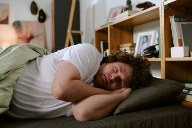 Sıcak ışık altında, rahat bir yatak odasında huzur içinde uyuyan bir adam. Etrafında raflar ve kişisel eşyalar, rahat ve rahat bir atmosfer yaratıyor.