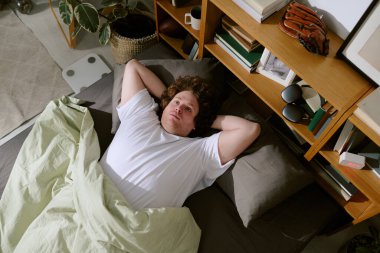 Yatağa uzanmış, elleri başının arkasında, kitaplarla ve bitkilerle dolu raflarla çevrili, yerde bir sepet ve yakınlarda beyaz bir halı ile.