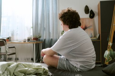 Güneşli bir odada yatakta oturup pencereden dışarı bakan bir adamın portresi. Etrafı kapalı bitkilerle ve minimalist mobilyalarla çevrili. Sakin ve huzurlu bir atmosfer yaratıyor.