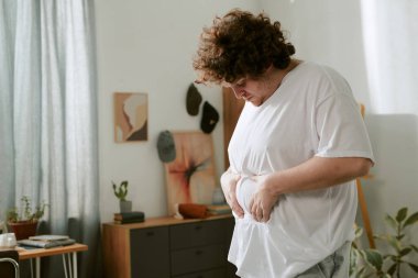 Yatak odasında üzerinde bitkiler ve duvar resimleri olan beyaz tişört giyen biri derin düşüncelere dalmış. Pencereden süzülen ışık, sakin atmosferi güçlendirir