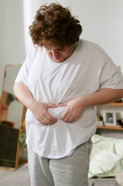 Konforlu yatak odasında duran kişi rahat ev kıyafetlerine bel bağlarken rahat ve rahat atmosferi yansıtıyor.